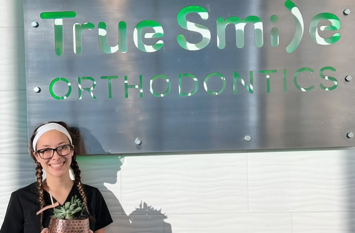 Young woman in black scrubs smiling while holding a succulent plant in front of the True Smile Orthodontics sign, promoting a welcoming atmosphere for cosmetic and orthodontic treatments.