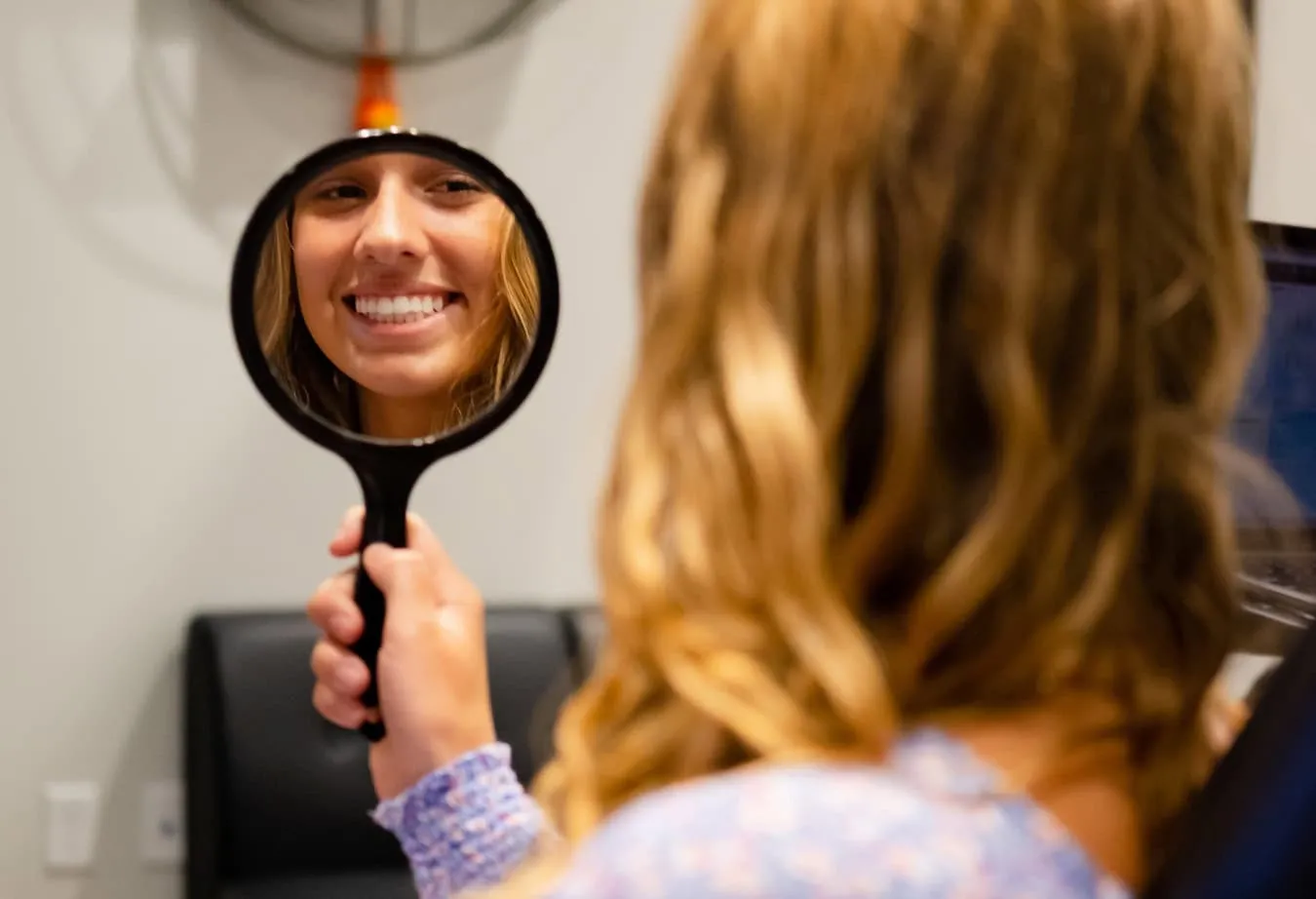 Smiling young woman looking in a handheld mirror, showcasing her teeth after orthodontic treatment, in a dental office setting.